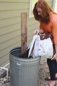 woman pouring a bag of potting soil in to a metal trash can with a board sitting in it